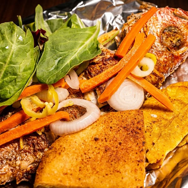 Fried fish with plantains, carrots, onions, and greens on a foil-lined plate.